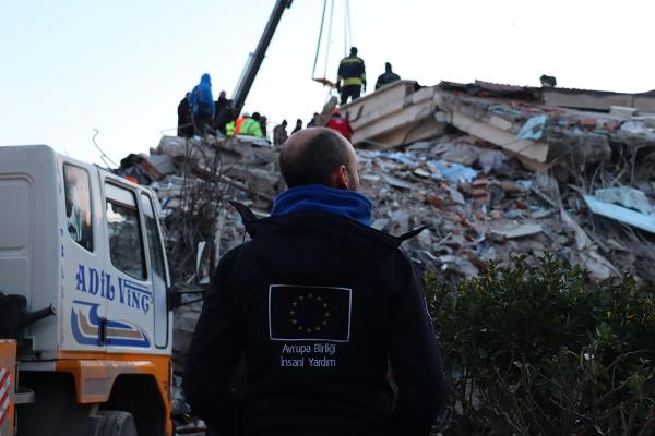 Aid worker, seen from the back with the EU flag logo on his vest, looking at the destructed buildings