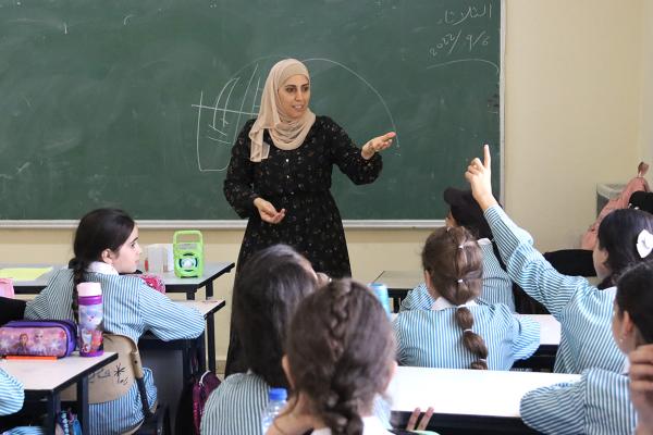 Teacher Anwar Bani Shamseh in front of a classroom, students raising hands to answer a question.
