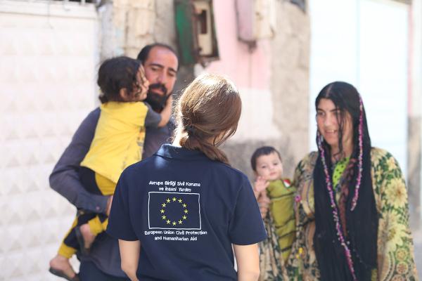 An aid worker, wearing a shirt with an EU flag, seen from the back talking with the family.