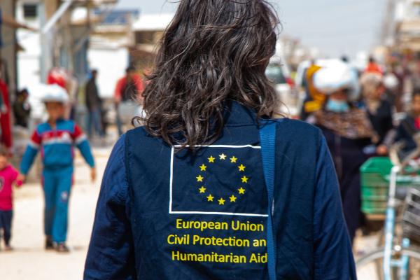 Aid worker in a street, seen from the back. A EU flag with text shown on the back of her jacket.