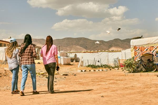 3 youngsters walking towards a settlement in a sandy area. In the background some mountains.