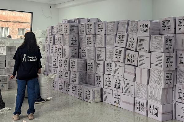 View of a big white room filled with boxes and blankets. Some people around it, in the background a window.