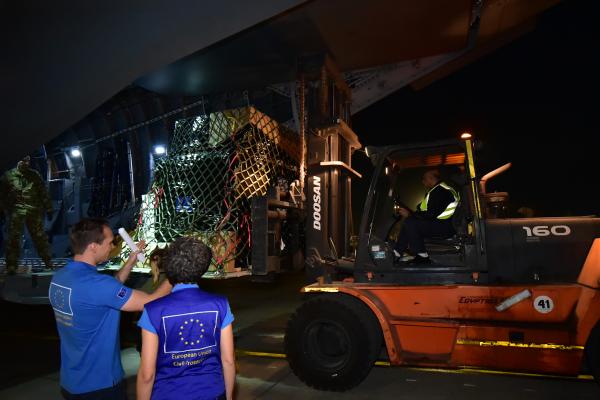 A forklift truck unloading goods form a plane in the dark. In front 2 EU Humanitarian Aid workers seen from the back.