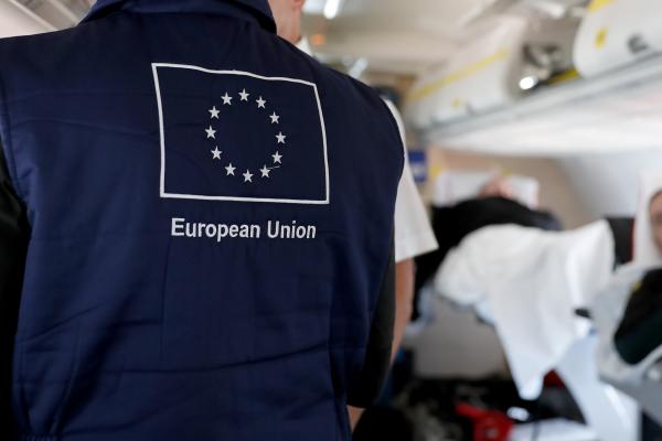 View inside a plane, a person wearing a vest with the EU flag. At the right a patient laying in a bed.