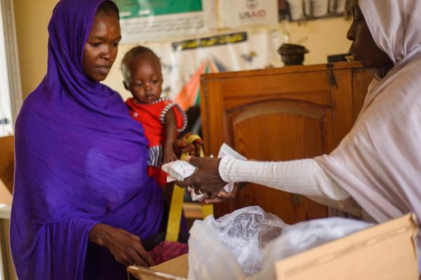 A woman with her daughter standing in a shop. They receive food sachets from a person at the right.