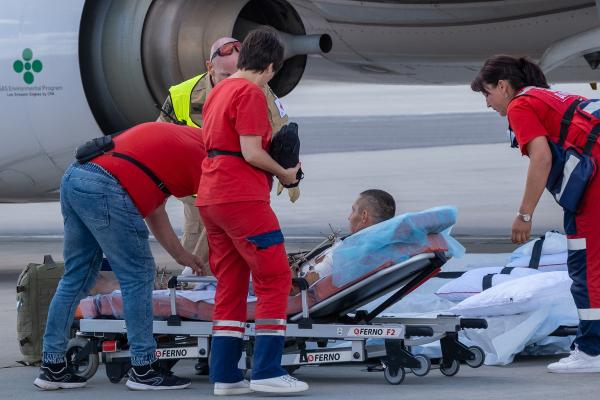Health workers taking care of a person on the tarmac of an airfield. In the background parts of an airplane are seen.